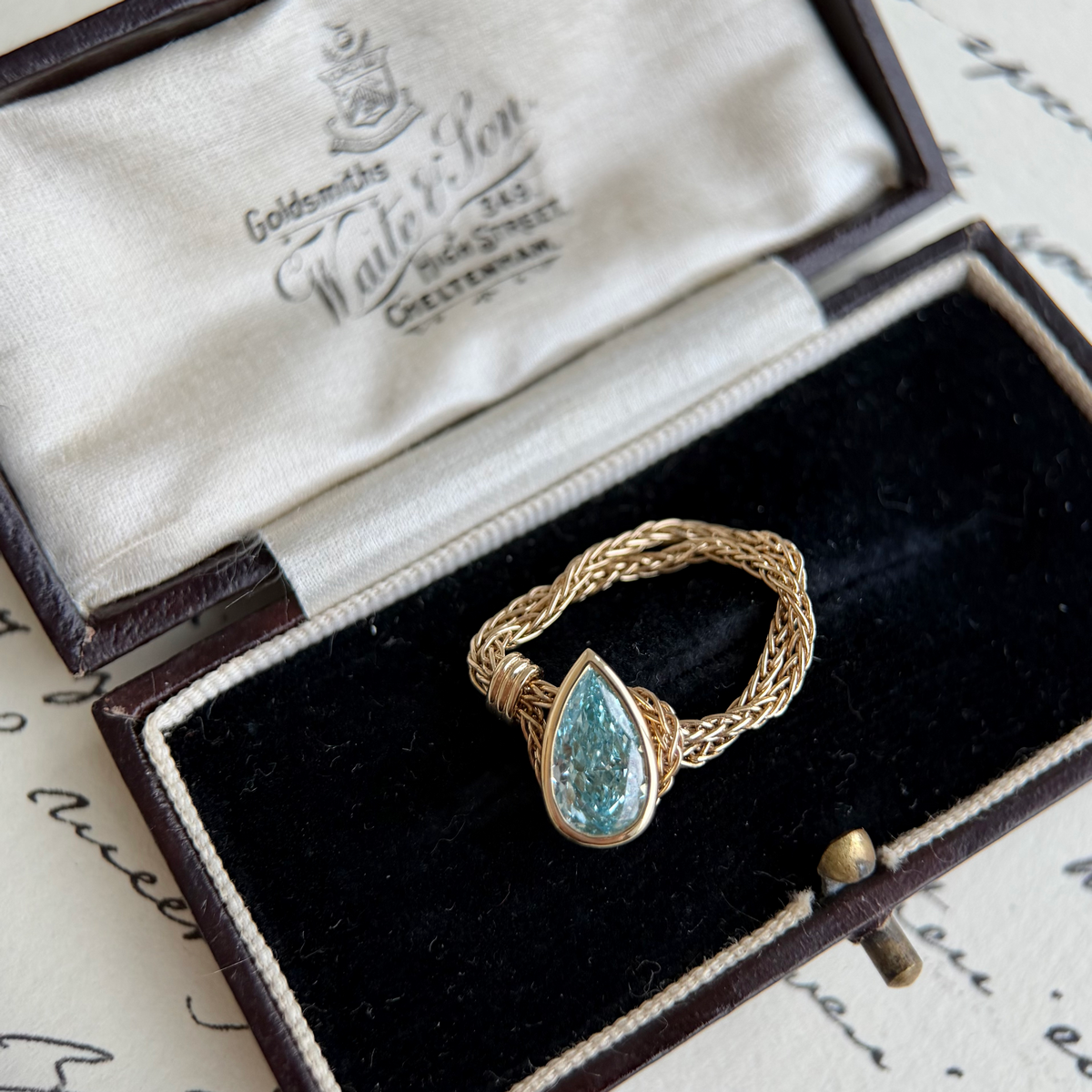 A gold ring with a teardrop-shaped blue-green stone, displayed inside an open vintage-style presentation box with a black velvet lining and cream satin interior.