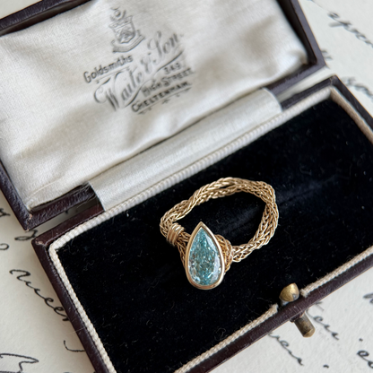 A gold ring with a teardrop-shaped blue-green stone, displayed inside an open vintage-style presentation box with a black velvet lining and cream satin interior.