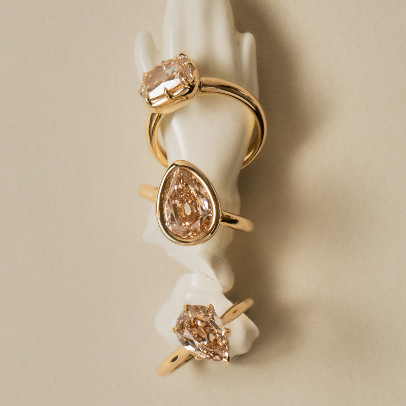 Three gold rings with large peach-colored diamonds displayed on a white sculptural hand stand against a warm beige background.
