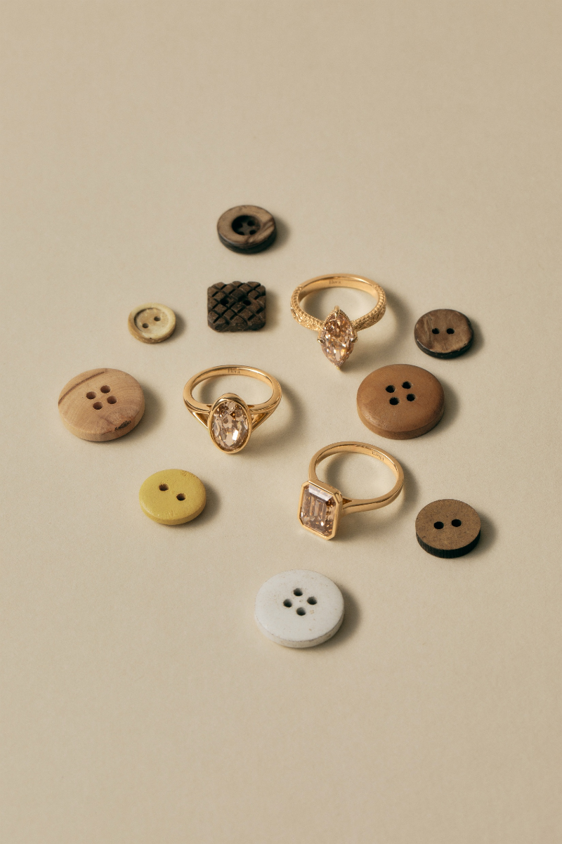 A styled flat-lay of mixed buttons and gold rings on a warm beige background. The collection includes wooden buttons in brown, yellow, and white, a textured black button, and several gold rings with clear stones.
