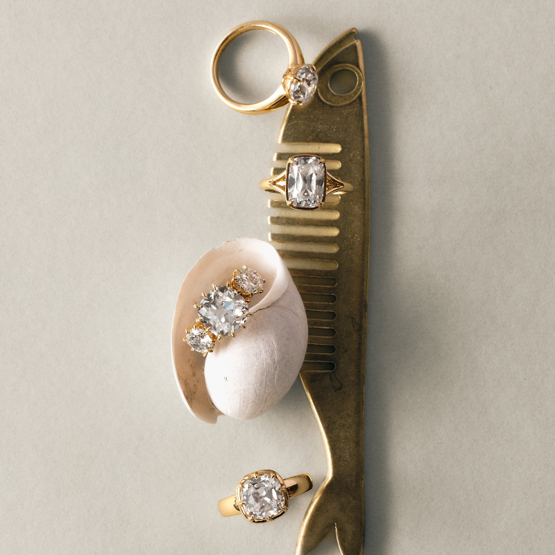 A gold-toned vintage comb with diamond accents, displayed on a neutral beige background alongside a small white shell and three rings featuring round-cut clear gemstones.