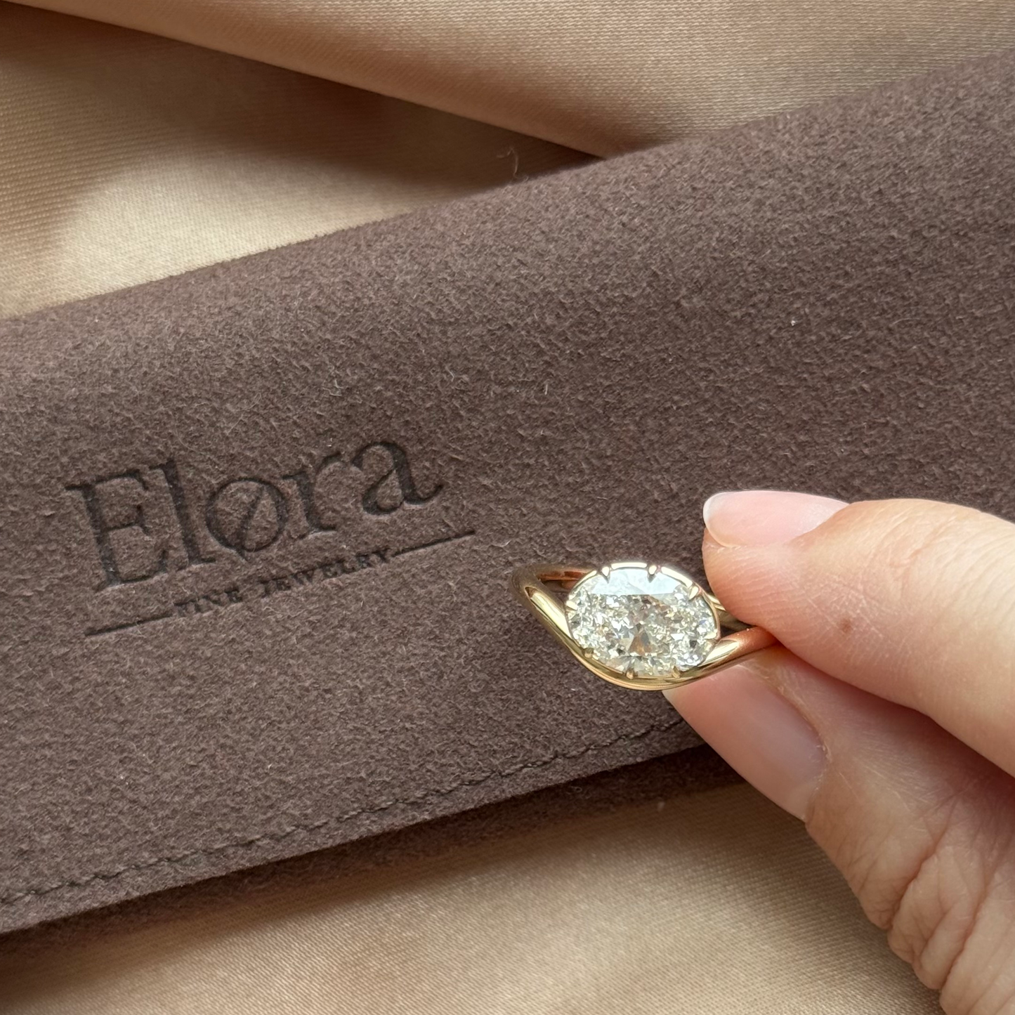 Wave-Oval Cut Lab Grown Diamond Engagement Ring held in hand with Elora Fine Jewelry branding in background.